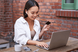 A smiling woman enters her credit card details into a laptop computer. There is a brick wall behind her. 