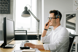 A smiling man sits on a desk with his hands on a computer keyboard. He is wearing glasses and a headset. There is a lamp behind him.